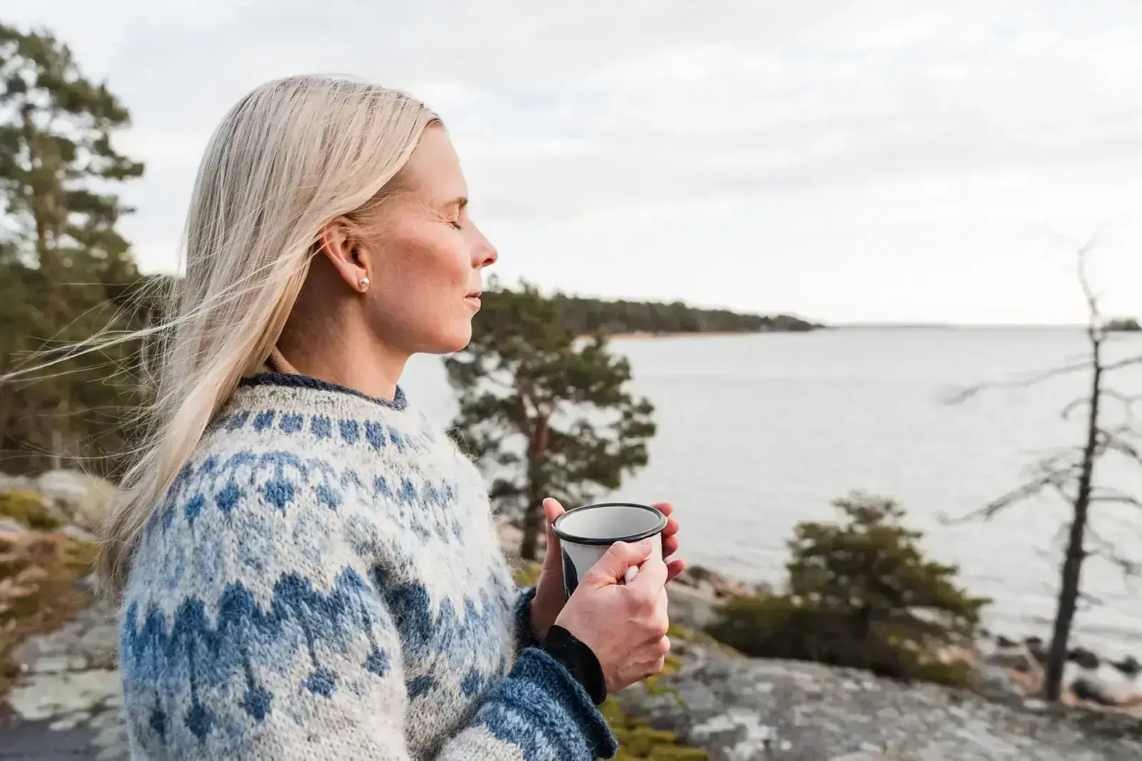 Vaalea nainen seisoo kalliolla kuppi kädessä ja katsoo merelle