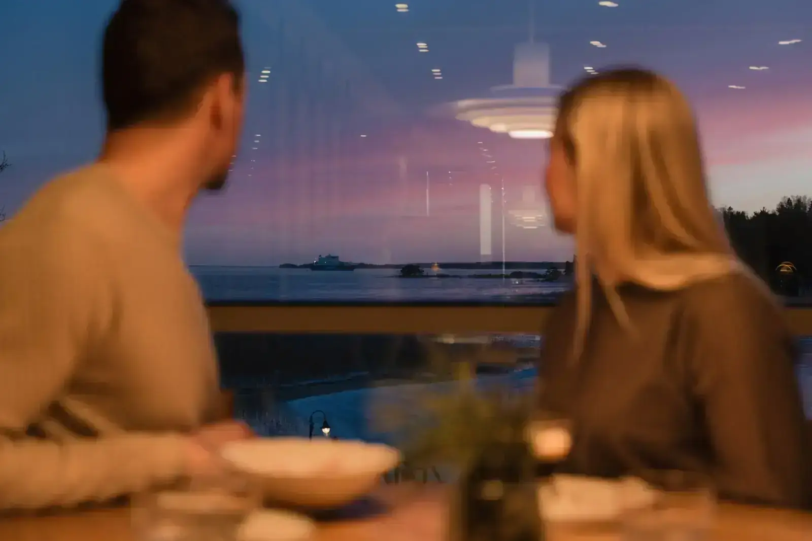 Kaksi ihmistä katsoo ravintolan ikkunasta iltaruskoa merelle